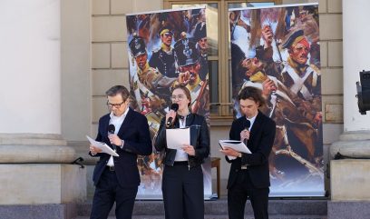 Trójka aktorów stoi przed wejściem do ratusza i czyta tekst.