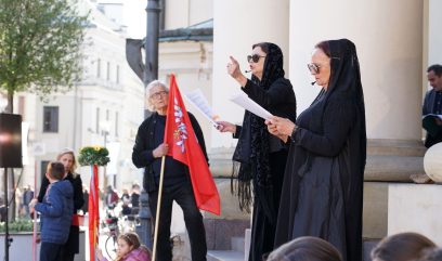 Dwie aktorki w czarnych szatach stoją przed wejściem do ratusza i czytają tekst. W tle Olgierd Łukaszewicz i dwójka dzieci trzymających chorągwie.