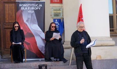 Olgierd Łukaszewicz czyta tekst. Za nim dwie aktorki w ciemnych szatach siedzą na krzesłach.