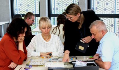 3 osoby siedzą przy stoliku. Nad nimi nachyla się instruktorka i tłumaczy zasady gry. Na stoliku leży rozłożona gra planszowa.