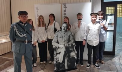 7 uczniów w mężczyzna w mundurze pozują z kartonowa figurą marszałka Piłsudskiego. Kilka osób przykłada sobie do twarzy sztuczne wąsy.