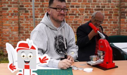 Mężczyzna siedzi przy maszynie do robienia przypinek. Przed nim kartonowy Archiwek.