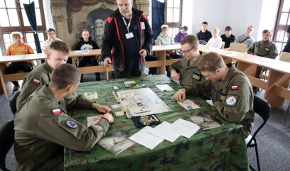 4 osoby siedzą przy stoliku i grają w grę pod okiem instruktora. W tle siedzi publiczność.