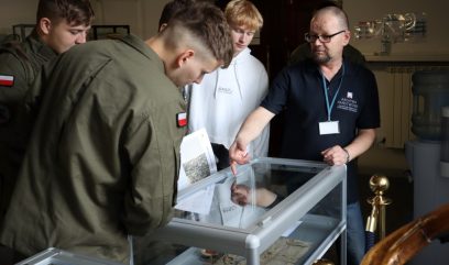 Goście oglądają materiały archiwalne wyłożone w szklanych gablotach.