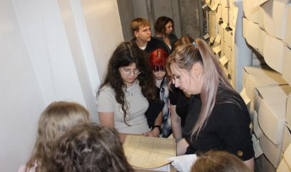 Magazyn archiwalny z regałami. Grupa dzieci otacza Archiwistkę, który trzyma otwartą księgę archiwalną.