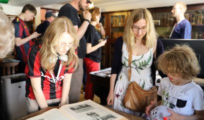 Grupa osób w bibliotece. 3 osoby pochylają się nad książką.