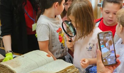 Dziewczynka ogląda przez lupę starą księgę. Z boku ktoś robi jej zdjęcie smartfonem.