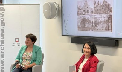 Dwie autorki siedzą na szarych fotelach. Prze nimi stolik. Na stoliku stoją 2 szklanki i 2 butelki wody. Za nimi na ścianie wisi ekran. Na ekranie wyświetlana jest prezentacja.