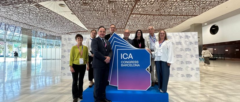 Naczelny Dyrektor wraz z kilkoma innymi osobami stoi przy banerze z logiem Międzynarodowego Kongresu Archiwów na niebieskim dywanie. Za nimi znajduje się ścianka z logiem Kongresu.