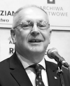 Doktor Maciej Janik w trakcie wygłaszania przemówienia. Przed nim znajduje się mikrofon a za nim - ścianka z logiem Archiwów Państwowych.