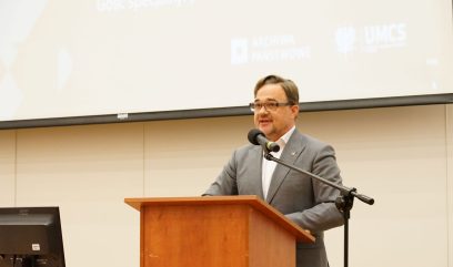 Aula wykładowa. Dyrektor Pietrzyk przemawia do mikrofonu. Przed nim siedzi publiczność.