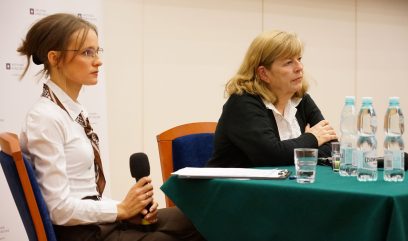 Aula wykładowa. Anne J. Gilliland siedzi na fotelu przy stoliku konferencyjnym i mówi do mikrofonu.