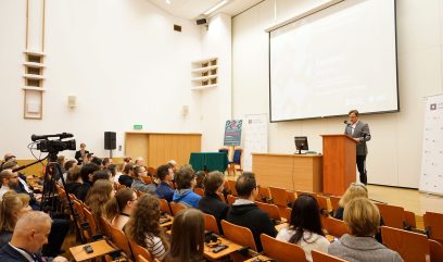 Aula wykładowa. Dyrektor Pietrzyk przemawia do mikrofonu. Przed nim siedzi publiczność.