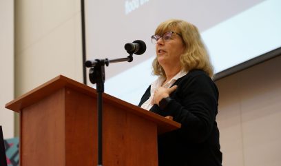 Aula wykładowa. Anne J. Gilliland na scenie przemawia do mikrofonu.