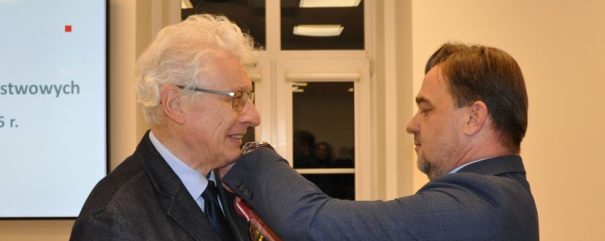 Mężczyzna w garniturze umieszcza medal z czerwono-białą wstążką na szyi starszego mężczyzny z białymi włosami i w okularach. Stoją w pomieszczeniu w pobliżu okna; ekran projekcyjny z tekstem jest częściowo widoczny po lewej stronie.