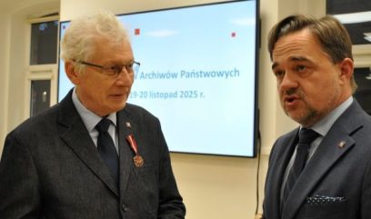 Dwóch mężczyzn w garniturach rozmawia w pomieszczeniu. Starszy mężczyzna po lewej ma siwe włosy, okulary i medal na klapie. Mężczyzna po prawej ma krótkie włosy i przystrzyżoną brodę. Za nimi znajduje się ekran z polskim tekstem i okno po lewej stronie.