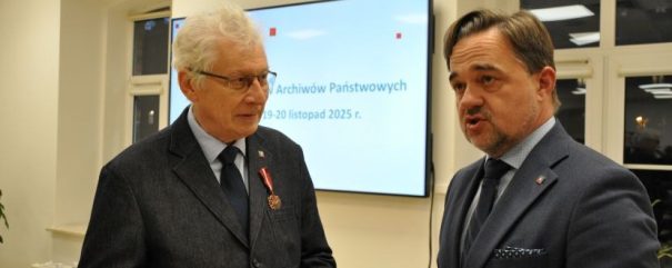 Dwóch mężczyzn w garniturach rozmawia w pomieszczeniu. Starszy mężczyzna po lewej ma siwe włosy, okulary i medal na klapie. Mężczyzna po prawej ma krótkie włosy i przystrzyżoną brodę. Za nimi znajduje się ekran z polskim tekstem i okno po lewej stronie.