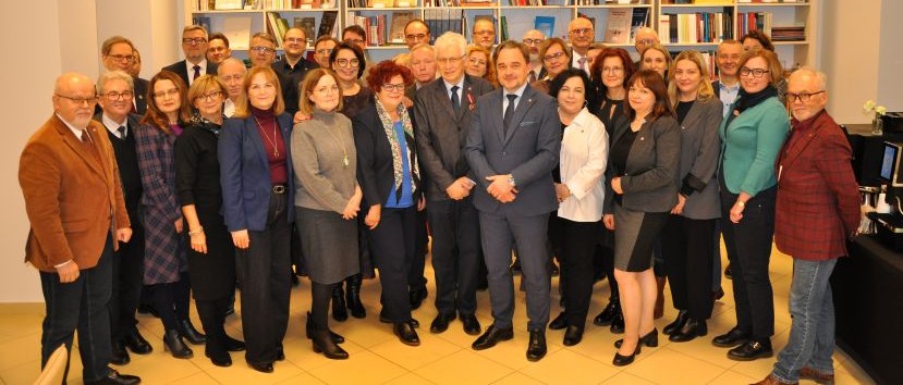 Duża grupa profesjonalnie ubranych dorosłych, mężczyzn i kobiet, stoi blisko siebie w pomieszczeniu przed półkami z książkami, uśmiechając się do kamery. Scenerią wydaje się być biuro lub biblioteka z półkami pełnymi książek w tle.