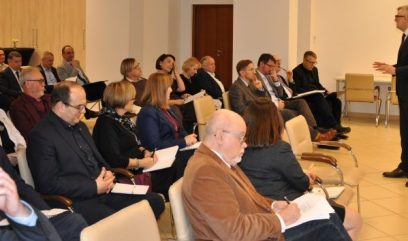 Mężczyzna w garniturze stoi i przemawia do publiczności siedzącej w rzędach krzeseł w jasno oświetlonym pomieszczeniu. Publiczność, składająca się z mężczyzn i kobiet, głównie w strojach biznesowych, słucha uważnie i robi notatki. Na pierwszym planie widać kilka pustych krzeseł.