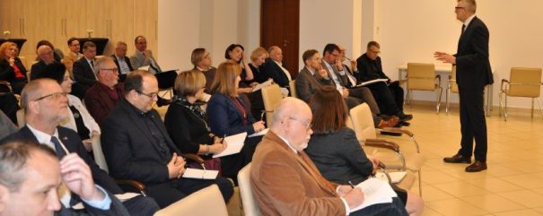 Mężczyzna w garniturze stoi i przemawia do publiczności siedzącej w rzędach krzeseł w jasno oświetlonym pomieszczeniu. Publiczność, składająca się z mężczyzn i kobiet, głównie w strojach biznesowych, słucha uważnie i robi notatki. Na pierwszym planie widać kilka pustych krzeseł.