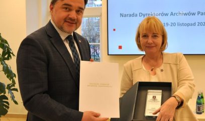 Mężczyzna w garniturze i kobieta w beżowej marynarce stoją w pomieszczeniu i oboje się uśmiechają. Mężczyzna trzyma teczkę, a kobieta otwarte pudełko z tabliczką. Za nimi znajduje się ekran z napisem Archiwa Państwowe i szczegółami konferencji.