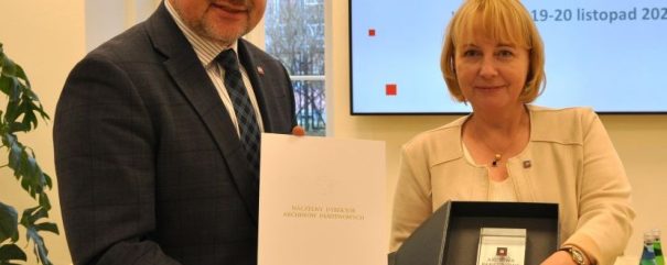 Mężczyzna w garniturze i kobieta w beżowej marynarce stoją w pomieszczeniu i oboje się uśmiechają. Mężczyzna trzyma teczkę, a kobieta otwarte pudełko z tabliczką. Za nimi znajduje się ekran z napisem Archiwa Państwowe i szczegółami konferencji.