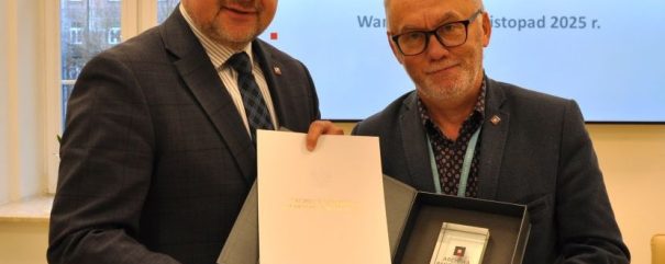 Dwóch mężczyzn w garniturach stoi w pomieszczeniu, uśmiechając się do kamery. Jeden z nich trzyma otwartą teczkę, a drugi odznaczenie. Za nimi znajduje się ekran telewizora wyświetlający polski tekst o spotkaniu archiwów państwowych zaplanowanym na listopad 2025 roku. W tle widać okno.