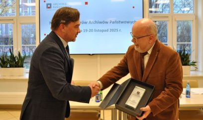 Dwóch mężczyzn w garniturach podaje sobie ręce; jeden wręcza drugiemu oprawiony certyfikat w czarnym pudełku. Stoją przed ekranem wyświetlającym polski tekst i datę, za nimi stół i butelki, a w tle duże okna ukazujące drzewa.