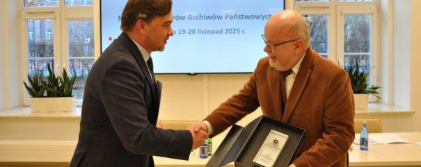 Dwóch mężczyzn w garniturach podaje sobie ręce; jeden wręcza drugiemu oprawiony certyfikat w czarnym pudełku. Stoją przed ekranem wyświetlającym polski tekst i datę, za nimi stół i butelki, a w tle duże okna ukazujące drzewa.