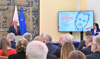 Mężczyzna przemawia na podium przed publicznością. Za nim znajdują się flagi Polski i UE. Po jego prawej stronie ekran wyświetla rysunek Aleksandra Kwaśniewskiego i tekst o dokumentach z jego prezydentury (1995-2005). Obok ekranu stoi kobieta.