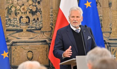 Starszy mężczyzna z siwymi włosami i brodą przemawia na podium. Za nim znajdują się flagi Polski i Unii Europejskiej oraz ozdobny gobelin. Ma na sobie granatowy garnitur, białą koszulę, czarny sweter i krawat, gestykuluje jedną ręką. Na pierwszym planie widoczne są głowy słuchaczy.