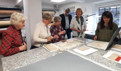 Pięć starszych kobiet i jeden mężczyzna stoją wokół nakrapianego blatu w jasnym pomieszczeniu, badając historyczne dokumenty. Niektóre dokumenty są otwarte, a grupa wydaje się być zaangażowana w dyskusję. W tle widoczne są półki, zwinięte materiały i duże okna.