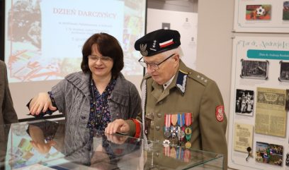 Starszy mężczyzna w mundurze wojskowym i czapce z wieloma medalami stoi obok uśmiechniętej kobiety. Oboje patrzą na dokumenty wystawione w szklanej gablocie. Za nimi znajdują się tablice informacyjne i ekran projektora z polskim tekstem.