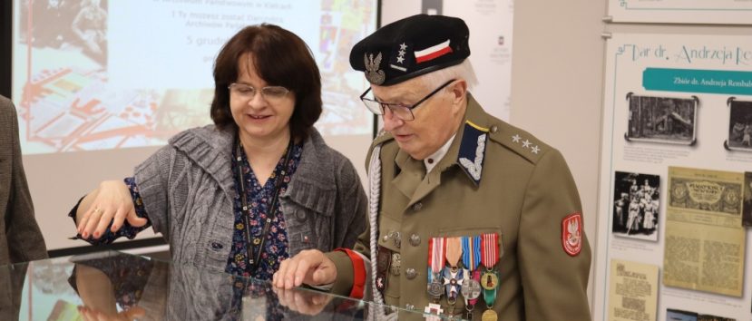 Starszy mężczyzna w mundurze wojskowym i czapce z wieloma medalami stoi obok uśmiechniętej kobiety. Oboje patrzą na dokumenty wystawione w szklanej gablocie. Za nimi znajdują się tablice informacyjne i ekran projektora z polskim tekstem.