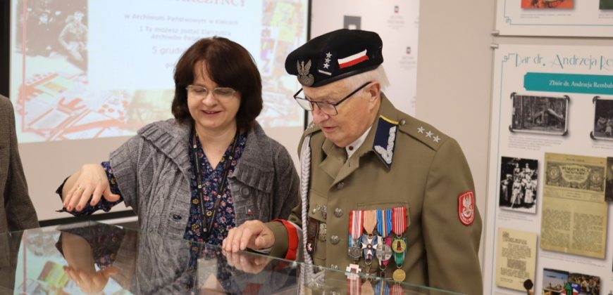 Starszy mężczyzna w mundurze wojskowym i czapce z wieloma medalami stoi obok uśmiechniętej kobiety. Oboje patrzą na dokumenty wystawione w szklanej gablocie. Za nimi znajdują się tablice informacyjne i ekran projektora z polskim tekstem.