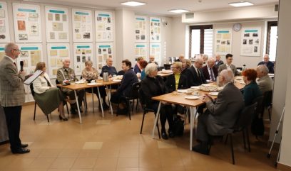 Grupa starszych osób siedzi przy stołach w jasno oświetlonym pokoju, słuchając stojącego mężczyzny, który mówi do mikrofonu. Na stołach leżą papiery i ciastka, a na ścianach za nimi wiszą stare dokumenty i zdjęcia.