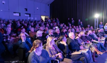Duża publiczność, składająca się głównie ze starszych osób, siedzi uważnie na wielopoziomowych siedzeniach w słabo oświetlonym audytorium. Ludzie noszą zimowe ubrania, a niektórzy trzymają gazety lub książki. Światło sceniczne świeci po prawej stronie, a ciemne zasłony zakrywają ścianę za nimi.