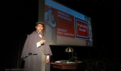 Osoba w stroju Sherlocka Holmesa, trzymająca fajkę, stoi przy biurku z zabytkowymi przedmiotami. Za nimi znajduje się duży ekran, na którym wyświetlany jest polski tekst o wydarzeniu wydawniczym i koszalińskich kryminałach. Tło jest ciemne, oświetlając scenę.