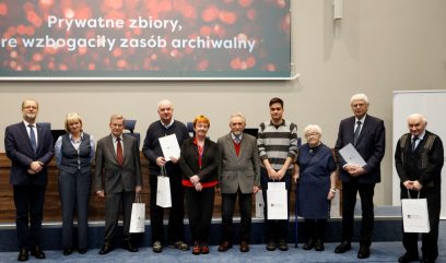 Grupa dziesięciu dorosłych stoi w rzędzie na scenie wyłożonej niebieskim dywanem, trzymając torby z prezentami i certyfikatami. Za nimi znajduje się ekran z czerwonymi światłami bokeh i polskim tekstem. Ludzie są ubrani formalnie i wydają się być na imprezie związanej z nagrodami lub uznaniem.