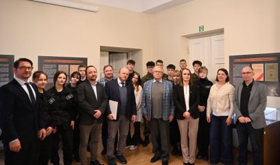 Grupa około 18 dorosłych i nastolatków stoi razem w dobrze oświetlonym pomieszczeniu z plakatami informacyjnymi na ścianach. Większość z nich ubrana jest w stroje biznesowe lub codzienne, a kilku w mundury. Stoją twarzą do kamery, pozując do zdjęcia grupowego.