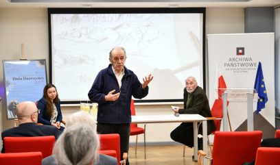 Starszy mężczyzna przemawia do publiczności w nowoczesnej sali konferencyjnej. Dwie osoby siedzą za stołem obok niego. Duży szkic jest wyświetlany na ekranie za nimi, a niebieska flaga UE stoi obok napisu Archiwa Państwowe. Kilka osób słucha z uwagą.