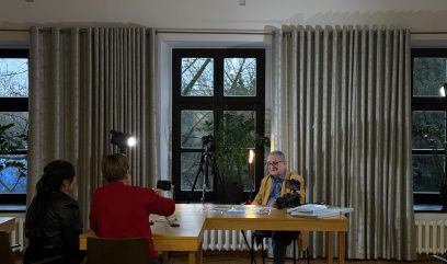 Trzy osoby znajdują się w dobrze oświetlonym pokoju z dużymi oknami i beżowymi zasłonami. Dwie kobiety siedzą przy stole naprzeciwko mężczyzny w okularach, który uśmiecha się i siedzi za kamerą i dokumentami. Statywy i światła są ustawione, sugerując wywiad lub nagrywanie.