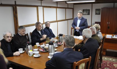 Grupa starszych dorosłych i jeden młodszy mężczyzna siedzą wokół drewnianego stołu z filiżankami do kawy, termosami, cukrem i przekąskami. Młodszy mężczyzna stoi i przemawia do grupy. Pokój jest skromny, z białymi panelami na ścianach, drewnianymi belkami i wiszącymi portretami.