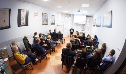 Grupa ludzi siedzi na krzesłach w małej sali konferencyjnej, naprzeciwko mężczyzny prowadzącego prezentację. Na białych ścianach wiszą plakaty i certyfikaty, a projektor wyświetla slajdy. Okna wpuszczają naturalne światło z tyłu pomieszczenia.