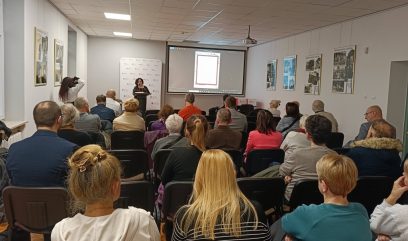 Kobieta stoi na podium, prowadząc prezentację dla publiczności siedzącej w rzędach w jasnym pomieszczeniu. Ekran projekcyjny z przodu wyświetla dokument. Ludzie są uważnie zwróceni do przodu. Fotografie i plakaty wiszą na białych ścianach wokół pokoju.