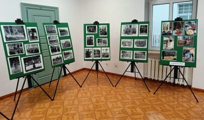 Pięć sztalug wyświetla zielone tablice z czarno-białymi i kolorowymi fotografiami w jasnym pomieszczeniu z podłogą wyłożoną kafelkami, białymi ścianami, zielonymi drzwiami i oknem. Obrazy wydają się przedstawiać historyczne wydarzenia i spotkania.