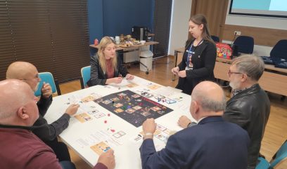 Sześć dorosłych osób siedzi przy stole i gra w złożoną grę planszową z kartami, kostkami i centralną planszą. Stojąca kobieta, prawdopodobnie moderator, zwraca się do grupy. Pokój ma nowoczesne meble, drewnianą podłogę i sprzęt do prezentacji w tle.