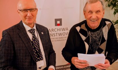 Dwóch starszych mężczyzn stoi uśmiechniętych w pomieszczeniu. Jeden nosi okulary, ciemny garnitur w kratę, białą koszulę i krawat w kropki. Drugi ma na sobie czarny sweter w beżowe wzory i trzyma dokumenty. Za nimi znajduje się baner z polskim tekstem i czerwono-czarnym logo.