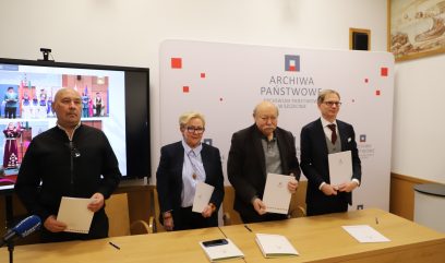 Cztery osoby stoją w rzędzie trzymając dokumenty przed znakiem Archiwa Państwowe w sali konferencyjnej. Mężczyzna po lewej stronie jest ubrany na czarno; pozostali są w strojach biznesowych. Telewizor po lewej stronie pokazuje połączenie wideo z kilkoma widocznymi uczestnikami.