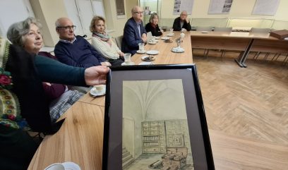 Grupa sześciu starszych osób siedzi wokół drewnianego stołu konferencyjnego z filiżankami i talerzami. Jedna z osób trzyma oprawiony rysunek pokoju z łukowatym sufitem, półkami, książkami i szafkami. Na ścianie za nimi wiszą historyczne plakaty.